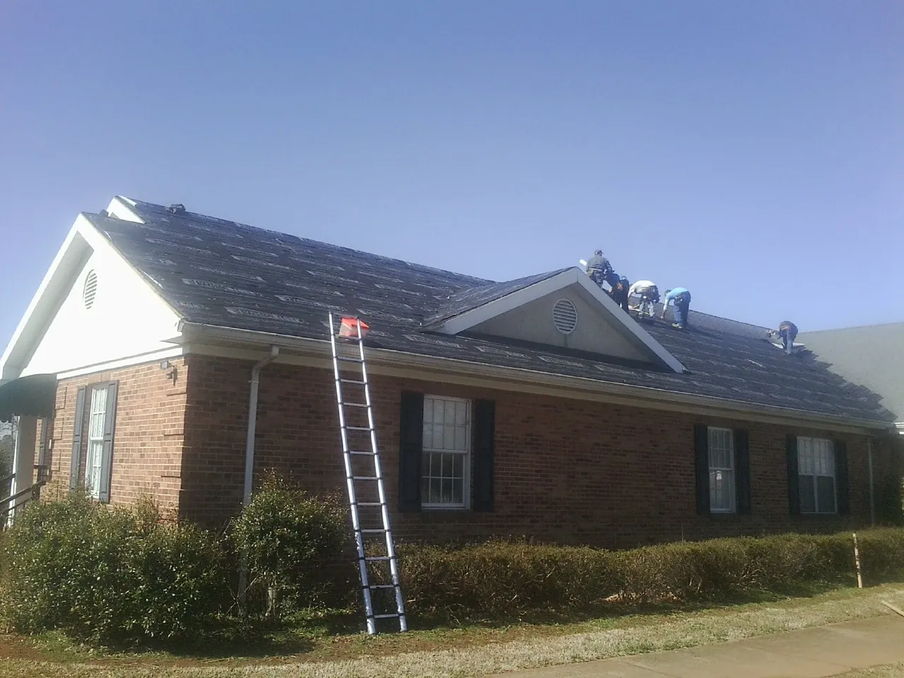 Crew tearing shingles off a brick ranch roof with a ladder set — Emergency Roof Repair work by our crew in Hall
