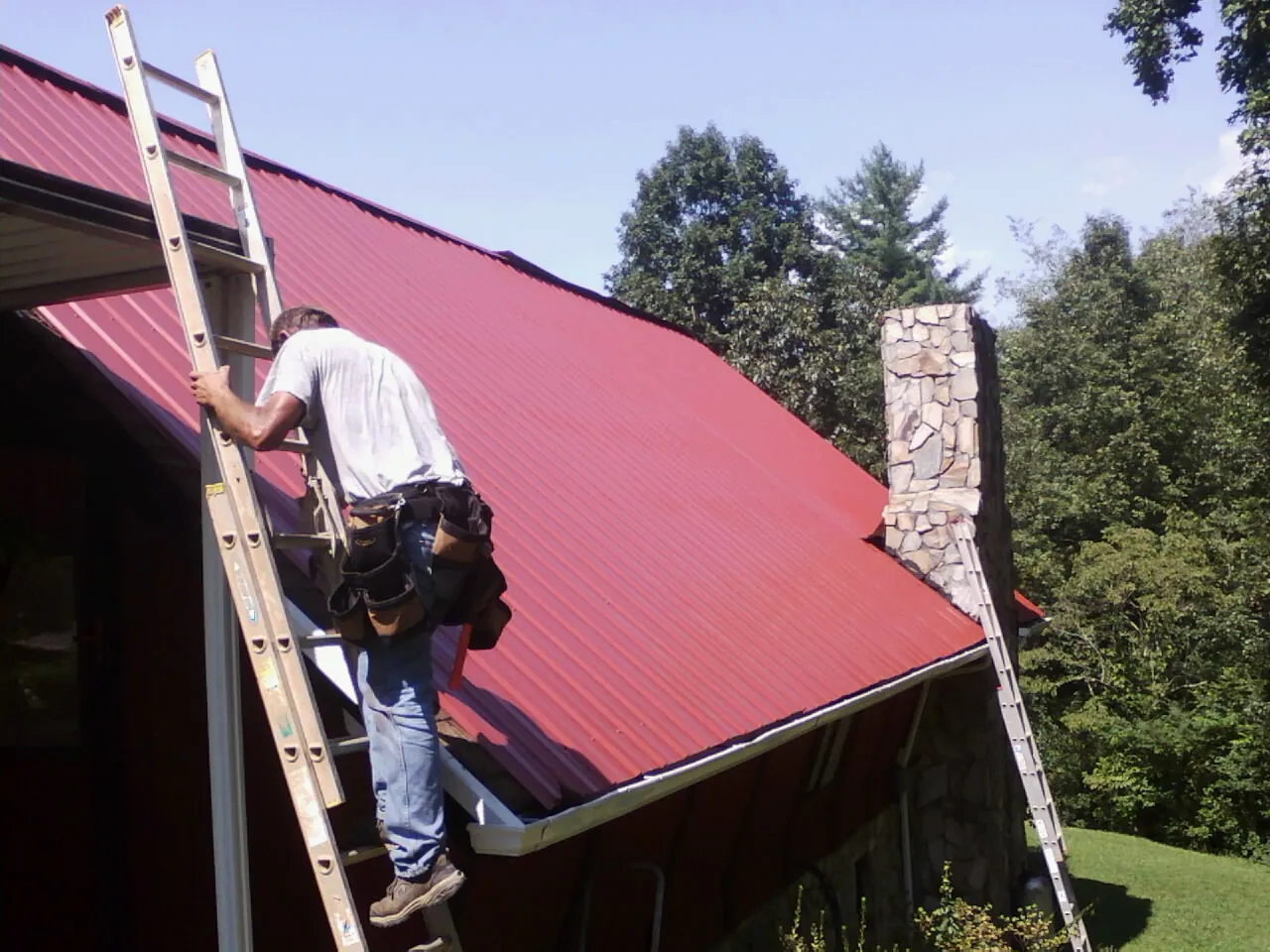 Roofer on a ladder beside a red metal roof and stone chimney — Soffit & Fascia Repair by Roofing Brothers in Hall
